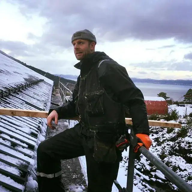 Bildet viser en mann med verktøy på tak i snøvær, klar for bygningsarbeid.